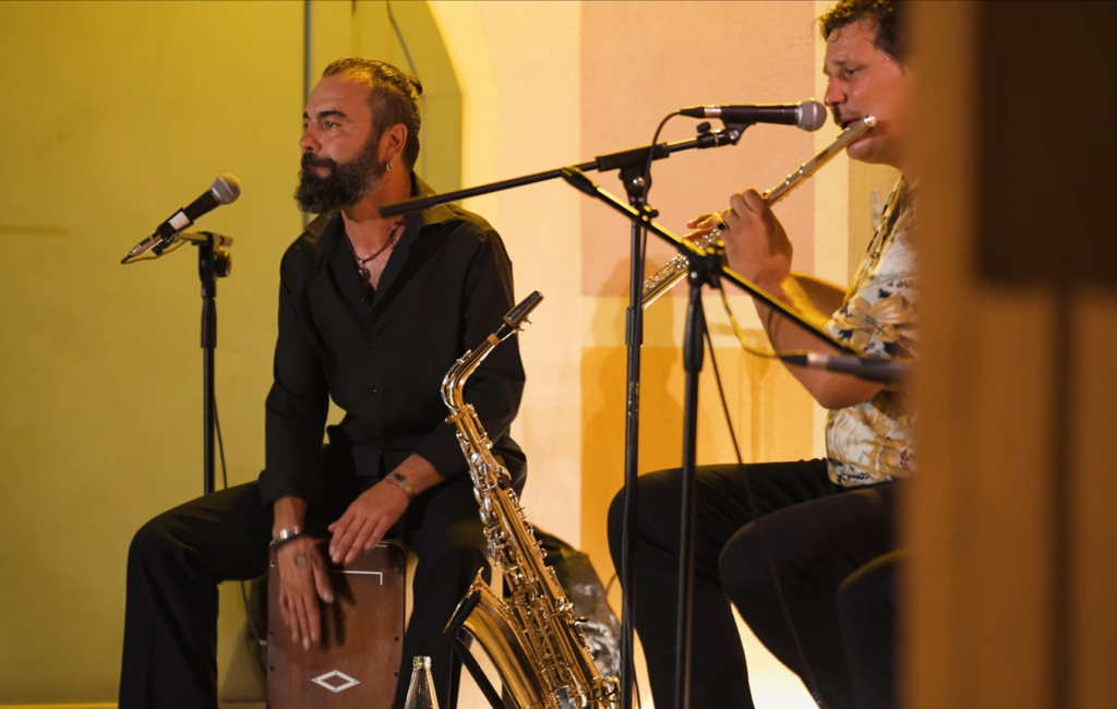 Two men on the stage together, one playing a flute and the other sitting on a drum which he's playing.